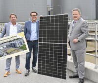 Drei Männer präsentieren an einem Klärbecken ein Solarmodul und eine Darstellung des geplanten Solarfaltdachs.