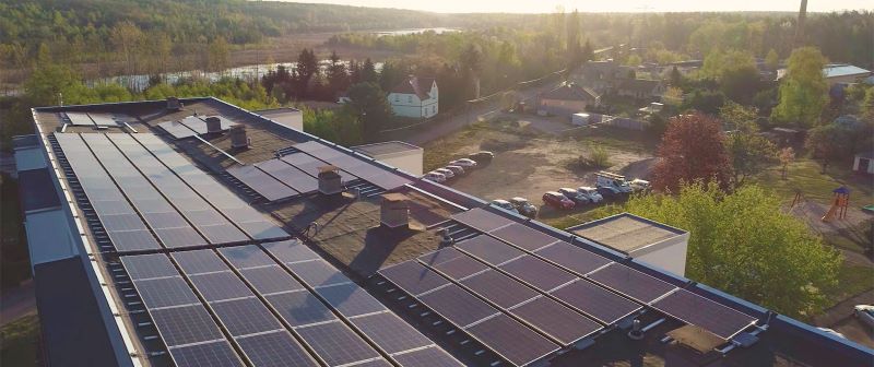 Blick auf ein Flachdach m,it Solarmodulen bei tioefstehender Sonne am Rande eines Wäldchens.