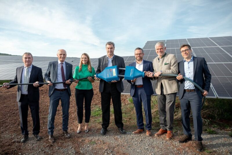 Sechs Männer und eine Frau halten ein dickes Kabel mit einem symbolischen Stecker - Inbetriebnahme am Solarpark Bundorf.