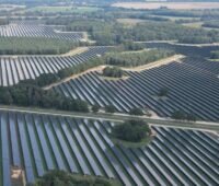 Luftaufnahme eines großflächigen Solarparks mit langen Reihen von Photovoltaik-Modulen zwischen Baumgruppen, Straßen und landwirtschaftlichen Flächen.