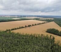 Luftbild einer Acker-Fläche, im Hintergrund Windenergie-Anlagen - hier soll der neue Solarpark entstehen.