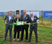 Vier Personen halten gemeinsam einen überdimensionierten grünen Stecker vor einer Freiflächensolaranlage.