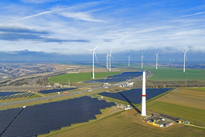 Ein Luftbild von schräg vorn mit Horizont: links oben Mitte der Tagebau Garzweiler, von links unten kommend die A44n mit Photovoltaikanlagen zu beiden Seiten sowie Windenergieanlagen dazwischen.