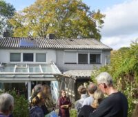 Menschengruppe von hinten im Garten eines Hauses mit kleiner PV-Anlage auf dem Dach: eine Solarparty.