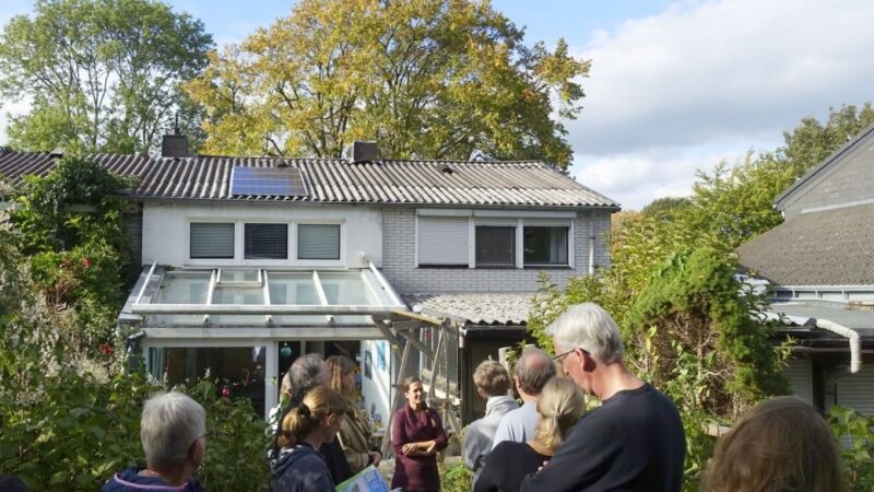 Menschengruppe von hinten im Garten eines Hauses mit kleiner PV-Anlage auf dem Dach: eine Solarparty.