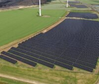 Luftaufnahme eines Solarparks entlang eines Autobahnabschnitts. Mehrere große Photovoltaikfelder liegen neben der Fahrbahn, im Hintergrund sind Windenergieanlagen und landwirtschaftliche Flächen zu sehen.