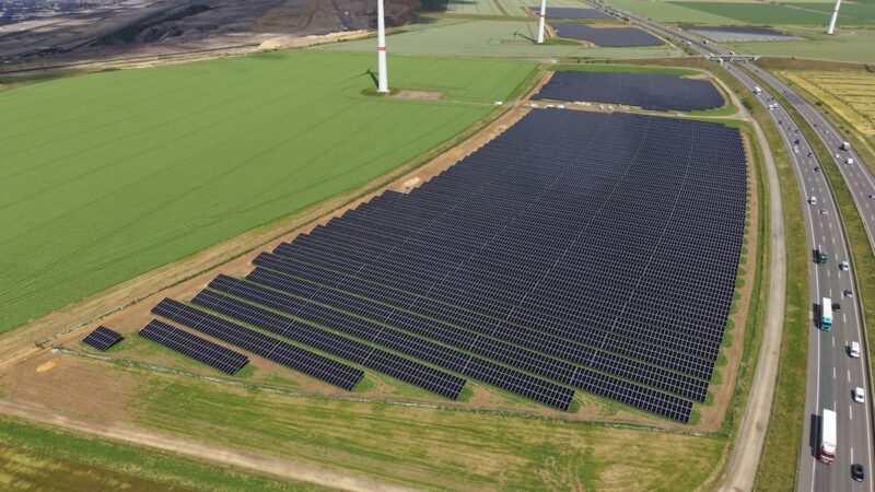 Luftaufnahme eines Solarparks entlang eines Autobahnabschnitts. Mehrere große Photovoltaikfelder liegen neben der Fahrbahn, im Hintergrund sind Windenergieanlagen und landwirtschaftliche Flächen zu sehen.