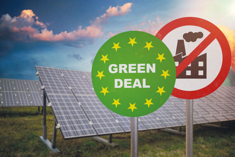 Symbolbild für EU-Solarstrategie: Schilder Green Deal und Abgase verboten vor einer Freiflächen-Solaranlage