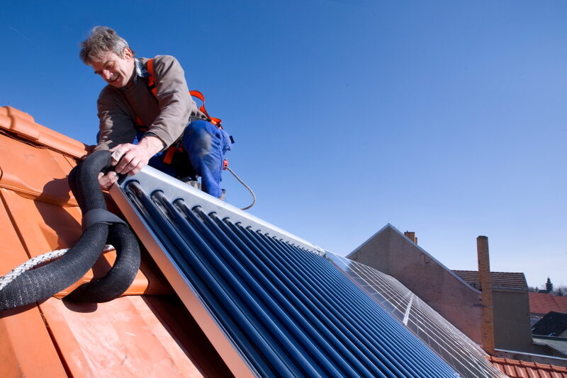 Ein Installateur montiert auf einem Spitzdach mit roten Schindeln einen blauen Solarwärmekollektor.