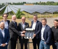 Sieben Personen stehen vor den aufgeständerten CPC-Vakuumröhrenkollektoren einer Solarthermieanlage und halten gemeinsam ein Kollektorelement als symbolisches Übergabeobjekt. Im Hintergrund sind die Kollektorreihen der Anlage sowie ein Industriegebäude zu sehen.