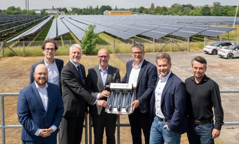 Sieben Personen stehen vor den aufgeständerten CPC-Vakuumröhrenkollektoren einer Solarthermieanlage und halten gemeinsam ein Kollektorelement als symbolisches Übergabeobjekt. Im Hintergrund sind die Kollektorreihen der Anlage sowie ein Industriegebäude zu sehen.