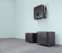 An einer Innenwand montierter Wechselrichter über zwei modularen Batteriespeichern von Solarwatt. Die Speichersysteme stehen auf dem Boden eines Technikraums und gehören zur neuen Heimspeicher-Generation Battery vision 2.0.