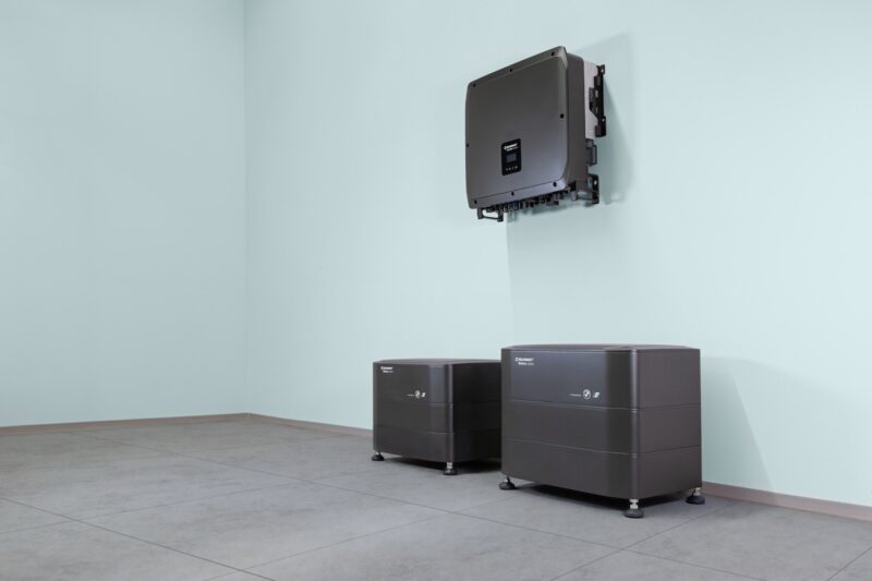 An einer Innenwand montierter Wechselrichter über zwei modularen Batteriespeichern von Solarwatt. Die Speichersysteme stehen auf dem Boden eines Technikraums und gehören zur neuen Heimspeicher-Generation Battery vision 2.0.
