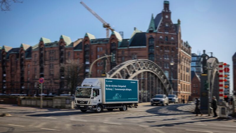 LKW mit Solarwatt-Schriftzug fährt über historische Brücke vor alten Backsteinhäusern - PV-Installation in Hamburg