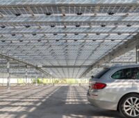 Blick unter eine Parkplatz-Überdachung mit halbtransparenten PV-Modulen, durch die Licht auf den Boden fällt, rechts steht ein Auto.