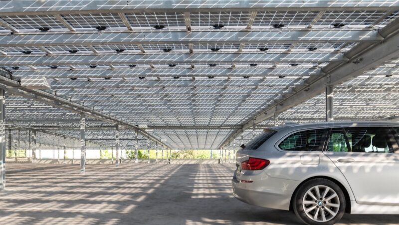 Blick unter eine Parkplatz-Überdachung mit halbtransparenten PV-Modulen, durch die Licht auf den Boden fällt, rechts steht ein Auto.