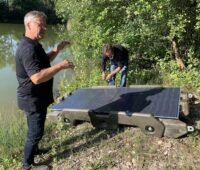 Zwei Männer und zwei Photovoltaik-Module auf einer Kunststoff-Konstruktion am Ufer - die Floating-PV-Anlage geht in den Test.