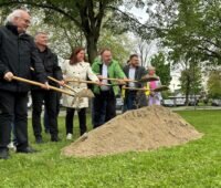 Männer und Frauen mit Spaten vor einem Haufen Sand - symbolischern Spatenstich für Ausbau von Wärmenetz mit Pelletkessel.