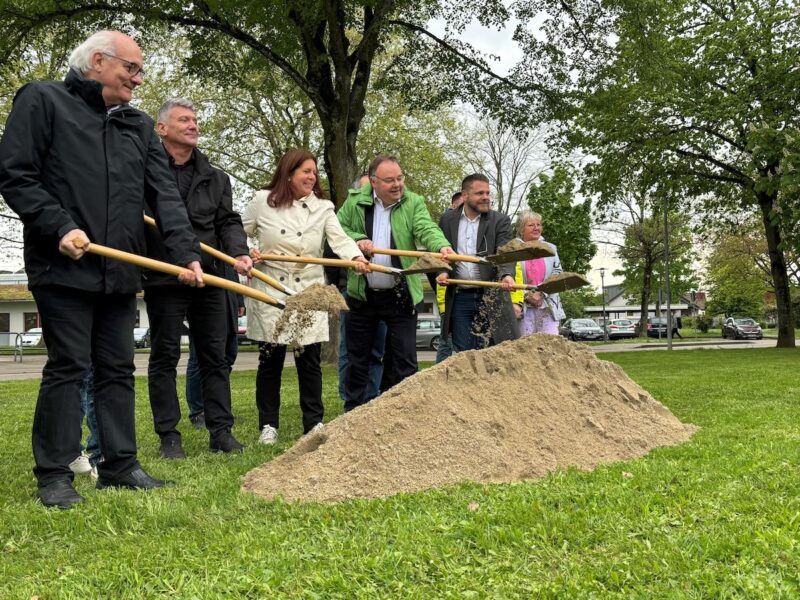 Männer und Frauen mit Spaten vor einem Haufen Sand - symbolischern Spatenstich für Ausbau von Wärmenetz mit Pelletkessel.