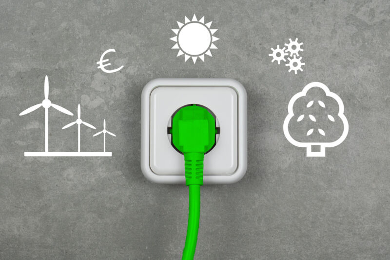 Graue Wand mit Symbolen für Windenergie, Sonne, Bioenergie und eine Steckdose mit einem grünen Stecker - Symbolbild für klimaneutrales Stromsystem