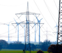 Hochspannungs-Strommasten im Vordergrund vor einem Windpark.