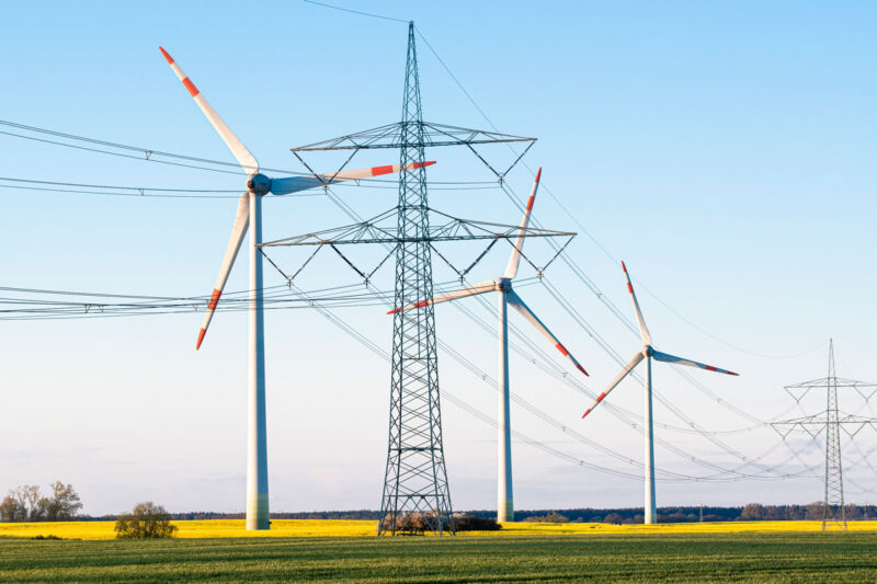 Hochspannungs-Strommast vor drei Windenergieanlagen. Symbolbild für Netzentgelte für Erzeugungsanlagen