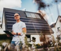 Ein Techniker mit einem Pad in der Hand vor einem Einfamilienhaus mit Dach-PV.