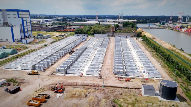 Luftbild von großem Batteriespeicherpark am Rande eines Kanals und eines Gewerbegebietes.