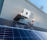 Zwei Monteure bringen an einer Wand den Sunny Tripower X 60-PV-Wechselrichter an. Er ist speziell für die Anforderungen gewerblicher PV-Anlagen entwickelt. Im Vordergrund befinden sich PV-Module.