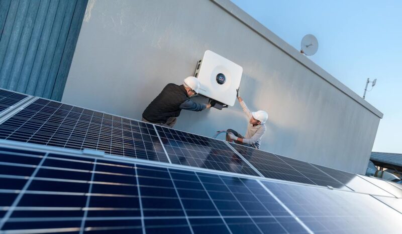 Zwei Monteure bringen an einer Wand den Sunny Tripower X 60-PV-Wechselrichter an. Er ist speziell für die Anforderungen gewerblicher PV-Anlagen entwickelt. Im Vordergrund befinden sich PV-Module.