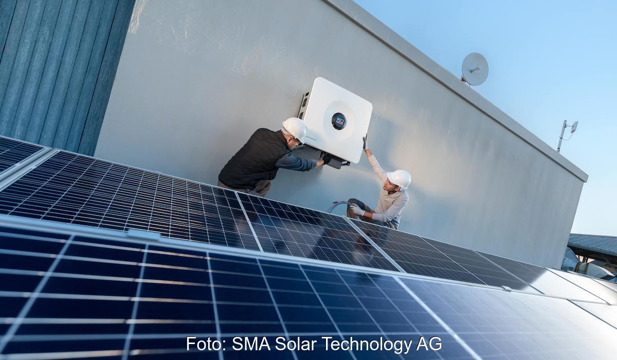 Zwei Monteure bringen an einer Wand den Sunny Tripower X 60-PV-Wechselrichter an. Er ist speziell für die Anforderungen gewerblicher PV-Anlagen entwickelt. Im Vordergrund befinden sich PV-Module.