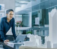 Eine Frau im Business-Outfit stützt sich in einer hell-modernen Büroumgebung auf einem Schreibtisch ab: TÜV Rheinland veröffentlicht Whitepaper mit neun Impulsen für eine umsetzungsorientierte kommunale Wärmeplanung.