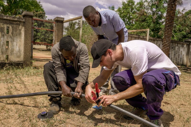Drei Männer arbeiten an einer Stromleitung in trockener buschigen Umgebung.