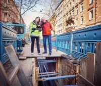 Das Foto zeigt zwei Personen stehend in einer Straßenschlucht mit mehrstöckigen Altbauten rechts und links. Unter ihren Füßen eine Baustelle, die Straße ist aufgerissen und legt Rohre frei.