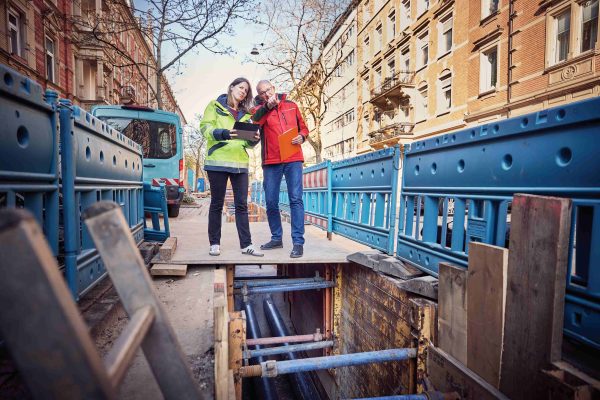 Das Foto zeigt zwei Personen stehend in einer Straßenschlucht mit mehrstöckigen Altbauten rechts und links. Unter ihren Füßen eine Baustelle, die Straße ist aufgerissen und legt Rohre frei.