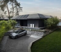 Auf dem Foto ist ein graues Einfamilienhaus mit PV-Anlage und zwei neuen, hellgrauen Tesla-Powerwall-3P zu sehen, davor, in der Zufahrt, ein grauer Tesla mit Wallbox. Das Haus ist umringt von Rasen, Bäumen und Sträuchern, im Hintergrund ein See durchs Grün und ein leicht diffuser Morgenhimmel.