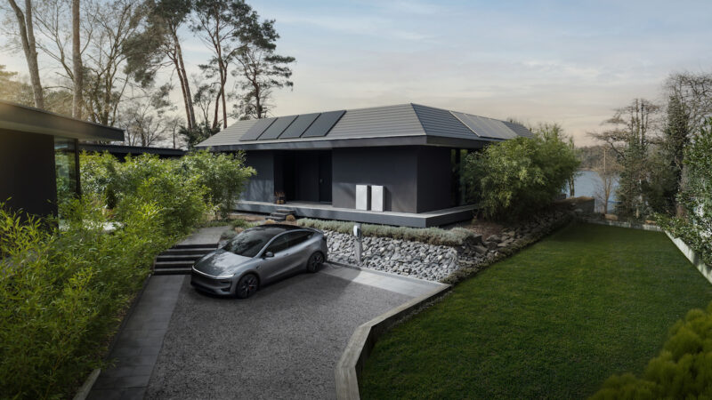 Auf dem Foto ist ein graues Einfamilienhaus mit PV-Anlage und zwei neuen, hellgrauen Tesla-Powerwall-3P zu sehen, davor, in der Zufahrt, ein grauer Tesla mit Wallbox. Das Haus ist umringt von Rasen, Bäumen und Sträuchern, im Hintergrund ein See durchs Grün und ein leicht diffuser Morgenhimmel.