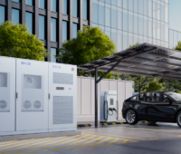 Das Rendering zeigt schräg links einen Batteriespeicher, rechts daneben ein schwarzer PKW, halb gezeigt, an einer überdachten Ladesäule, im Hintergrund Bäume und eine Glasfassade eines Hochhauses.