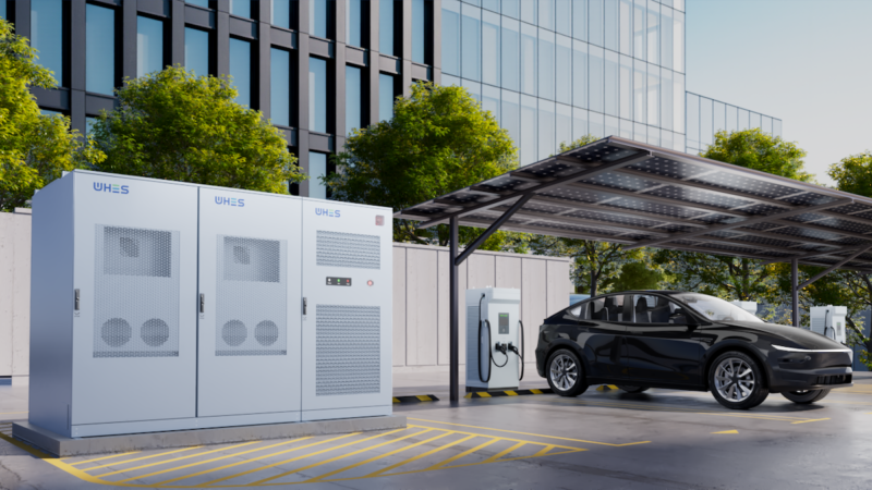 Das Rendering zeigt schräg links einen Batteriespeicher, rechts daneben ein schwarzer PKW, halb gezeigt, an einer überdachten Ladesäule, im Hintergrund Bäume und eine Glasfassade eines Hochhauses.