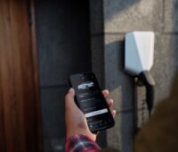 Hand hält Smartphone mit offener App zum Laden eines E-Autos - Wallbox im Hintergrund.
