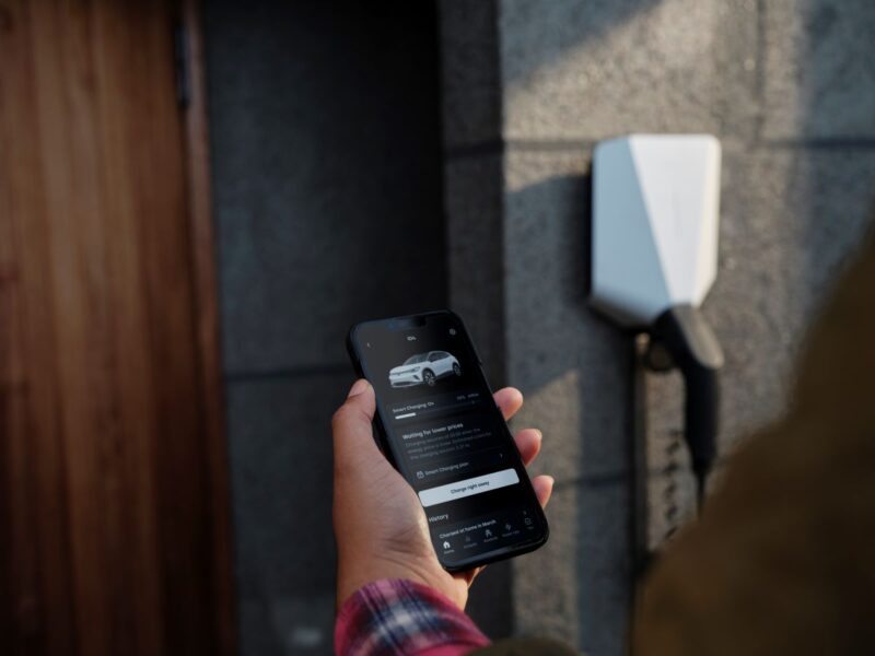 Hand hält Smartphone mit offener App zum Laden eines E-Autos - Wallbox im Hintergrund.