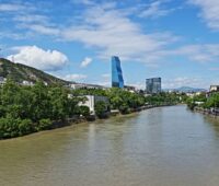 Ein breiter Fluss durchfließt die Stadt Tiflis: an den Ufern grüne Vegetation, Häuser und Hochhäuser im Hintergrund.