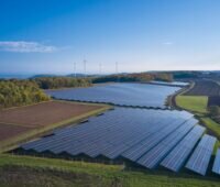 Luftaufnahme eines Freiflächensolarparks neben Äckern unter blauem Himmel.