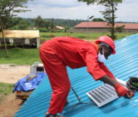 Ein Mann im roten Overall installiert auf einem Wellblechdach in Uganda ein Solarmodul.