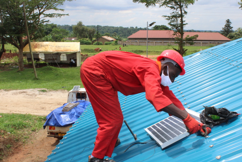 Ein Mann im roten Overall installiert auf einem Wellblechdach in Uganda ein Solarmodul.