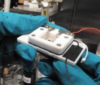 Zu sehen ist die Messzelle für die elektrochemische Rastermikroskopie, in mit blauen Händen bedeckten Händen.