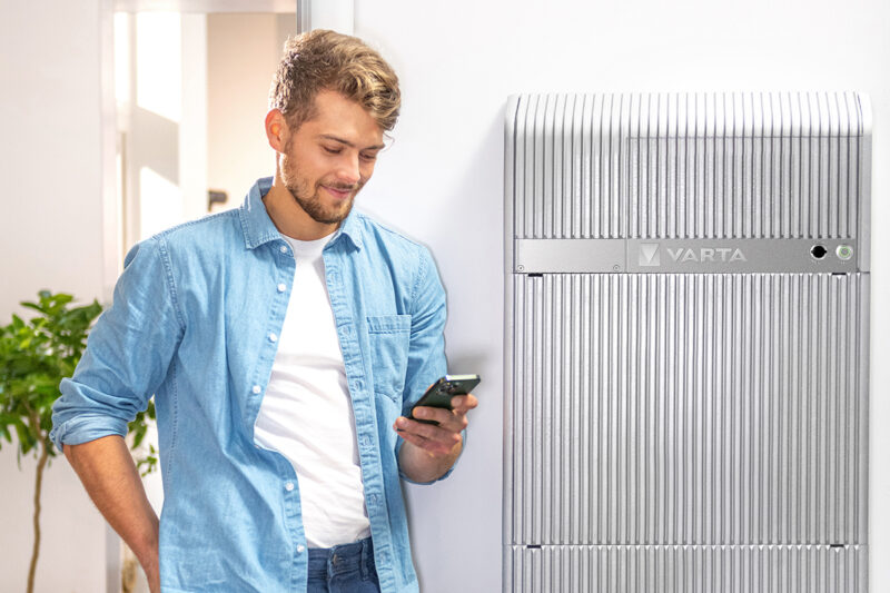 Blonder junger Mann mit Bart steht vor einer Varta Wall und schaut auf sein Smartphone.