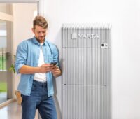 Blonder junger Mann mit Bart steht vor einer Varta Wall und schaut auf sein Smartphone.