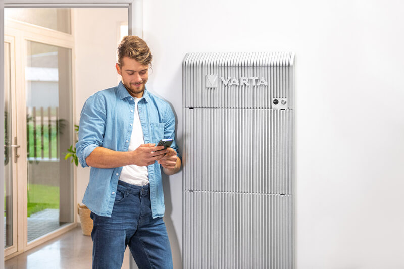 Blonder junger Mann mit Bart steht vor einer Varta Wall und schaut auf sein Smartphone.
