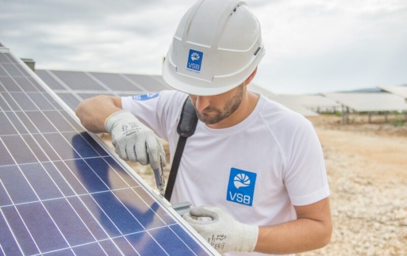Mann mit VSB-Logo montiert Photovoltaik-Modul auf einer Freifläche.